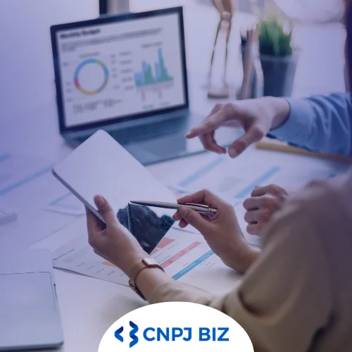 Relatório de Produtividade - Notebook aberto com dashboard na tela. Na mesa em que ele está há outros relatórios impressos e há mãos de pessoas com caneta e um tablet.